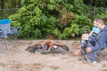 man, child, and dog sitting chair on sand next to fire