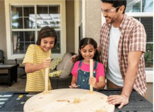 man and girls placing round sticks into holes on wooden board