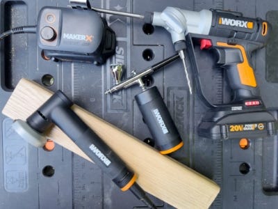 wooden block underneath rotary grinder tool, air brush, and drill on top of worx worktable