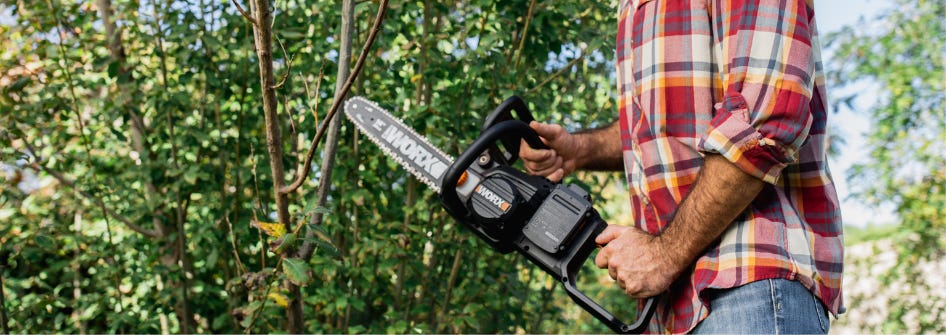 man holding worx cordless chainsaw in front of tree with green leaves