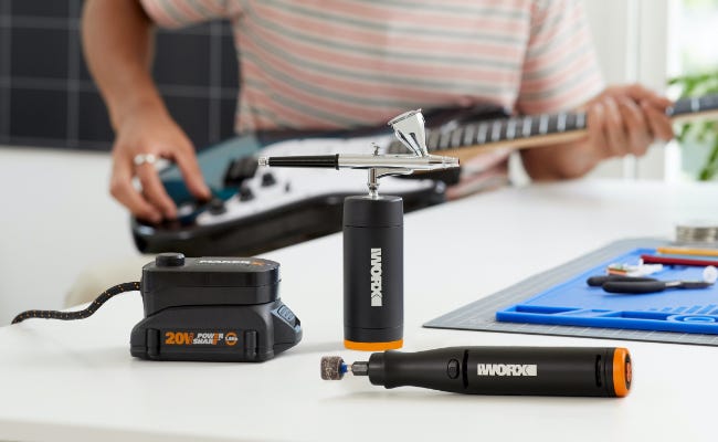 makerx rotary tool, airbrush and portable power source on a white table with a person playing a blue and white guitar in the background