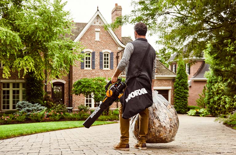 man holding bag of leaves with worx trivac and leaf collection bag in driveway in front of brick house linking to worx trivac product page