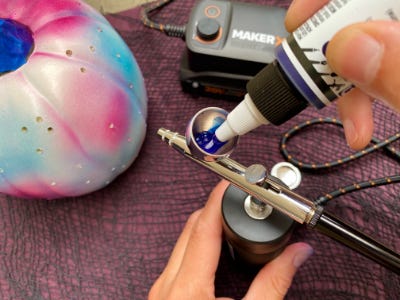 Pink, blue, and white pumpkin behind blue paint being filled into airbrush 