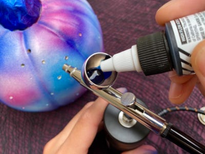 Pink, blue, black, and white pumpkin behind blue paint being filled into airbrush 