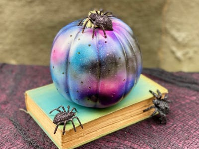 Galaxy color looking pumpkin sitting on top of book with two spiders near by