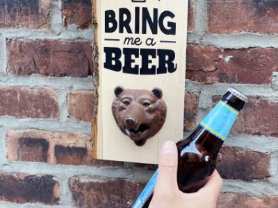 drink being held up next to bear bottle opener hanging on brick wall