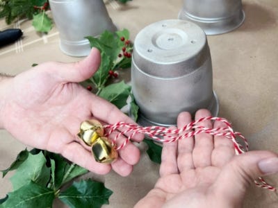 hands holding golden bells on red and white string in front of 3 silver pots