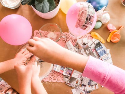 Hands adding stripped newspaper into flour water bowl mix with pink and yellow ballons behind