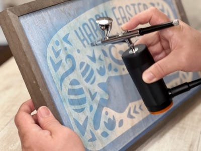 person using the air brush to paint their stencil over the wooden sign