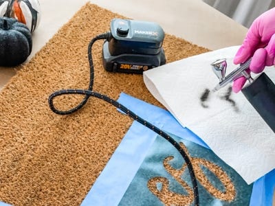 using airbrush to paint the words onto doormat with towel in background