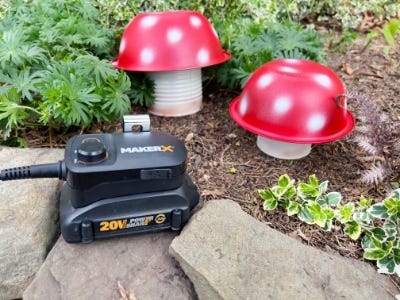 worx makerx charger and battery next to two red and white mushroom cans planted in dirt