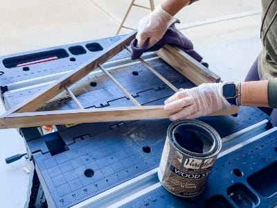 image of a person wearing gloves applying stain to the wooden tree on top the work table with the can of stain next to it 