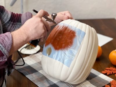 air brushing white pumpkin with stencil applied with hand holding it