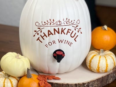 white pumpkin wine dispenser with thankful for wine and smaller pumpkins next to it on wooden plate