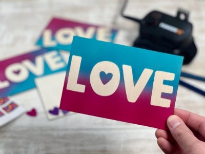 finished postcards in the background with the makerx hub and a person holding a finished postcard that is half blue and half pink with white letters in the foreground