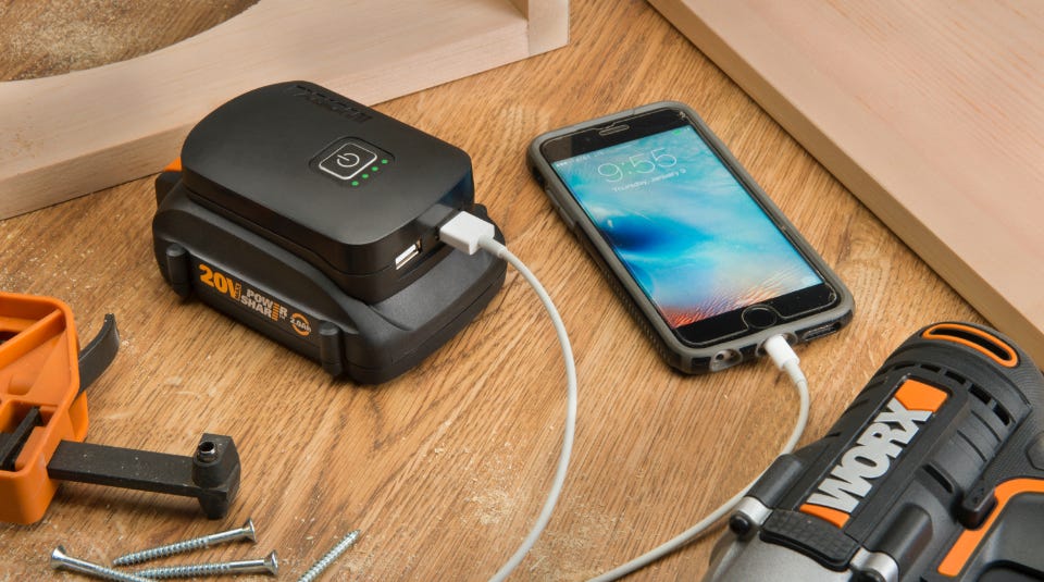 charger on the desk with a battery attached and a phone plugged into the usb port and a worx drill next to it