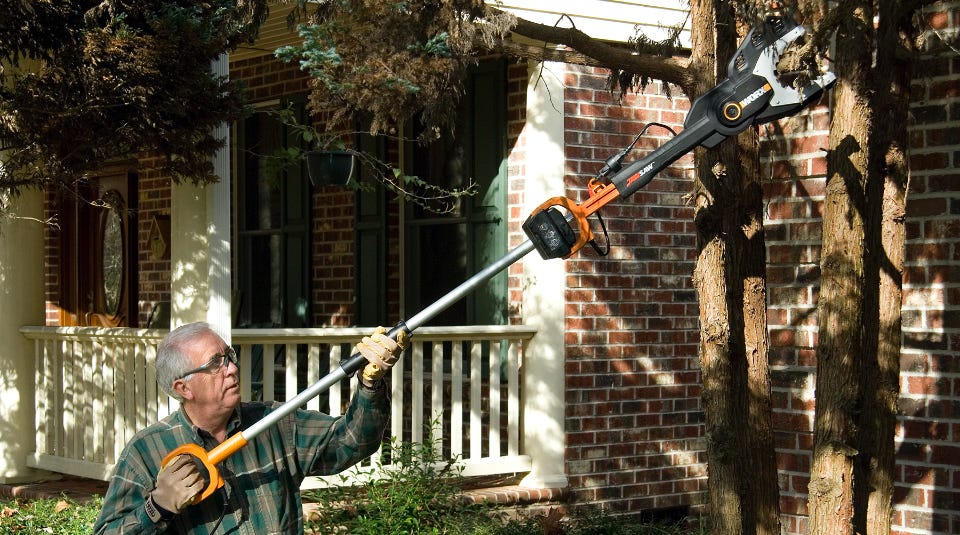 image of a man using the extension pole with a chainsaw connected to cut a branch off of a tree from in front of a house